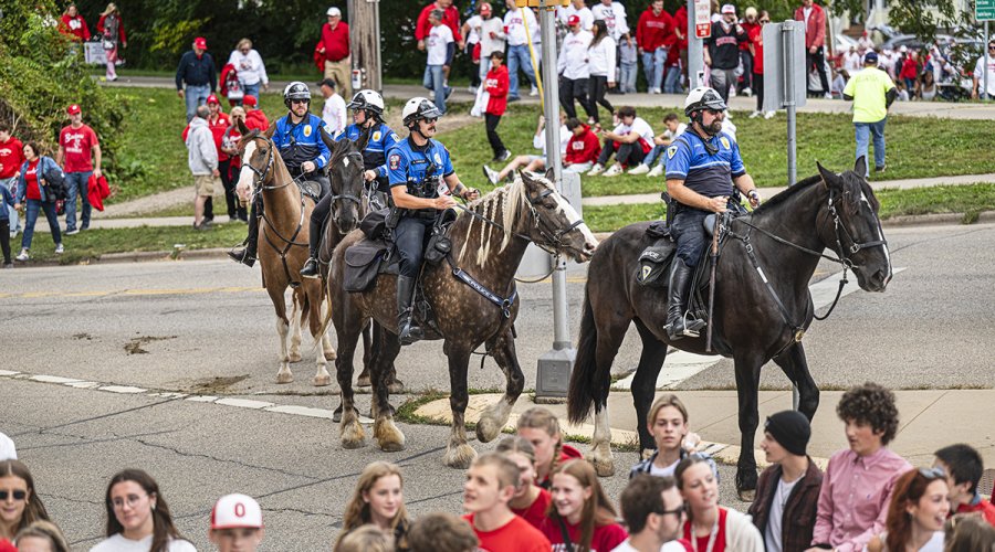 Four mounted police officers ride horses along a street during an event with a crowd of people wearing red and white in the background.