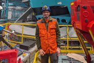 Keith Bechtol, wearing an orange safety vest and blue hard hat, stands in front of a large industrial machine surrounded by equipment and structures inside a spacious facility.