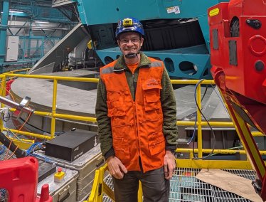 Keith Bechtol, wearing an orange safety vest and blue hard hat, stands in front of a large industrial machine surrounded by equipment and structures inside a spacious facility.