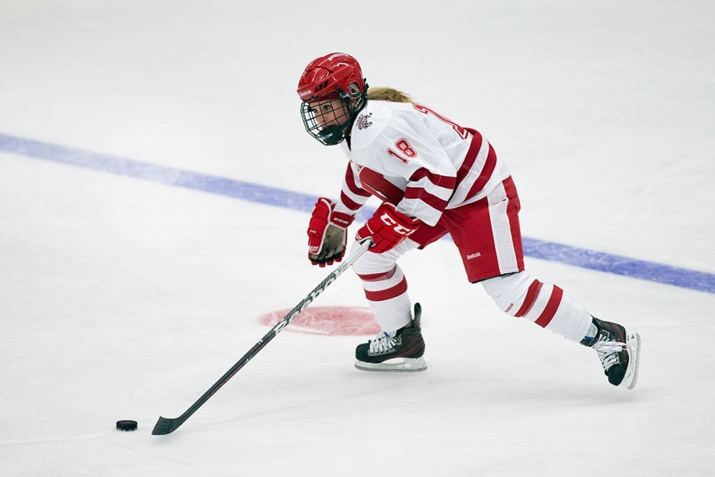 A hockey player in a UW red and white uniform skates forward on the ice while controlling the puck with the stick.