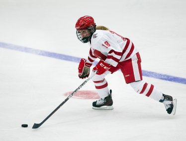 A hockey player in a UW red and white uniform skates forward on the ice while controlling the puck with the stick.