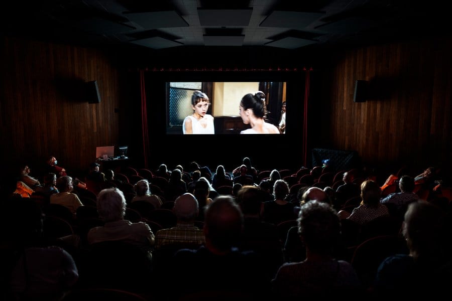 An audience sits in a dark theater watching a movie projected on a large screen.