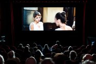 An audience sits in a dark theater watching a movie projected on a large screen.