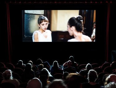 An audience sits in a dark theater watching a movie projected on a large screen.