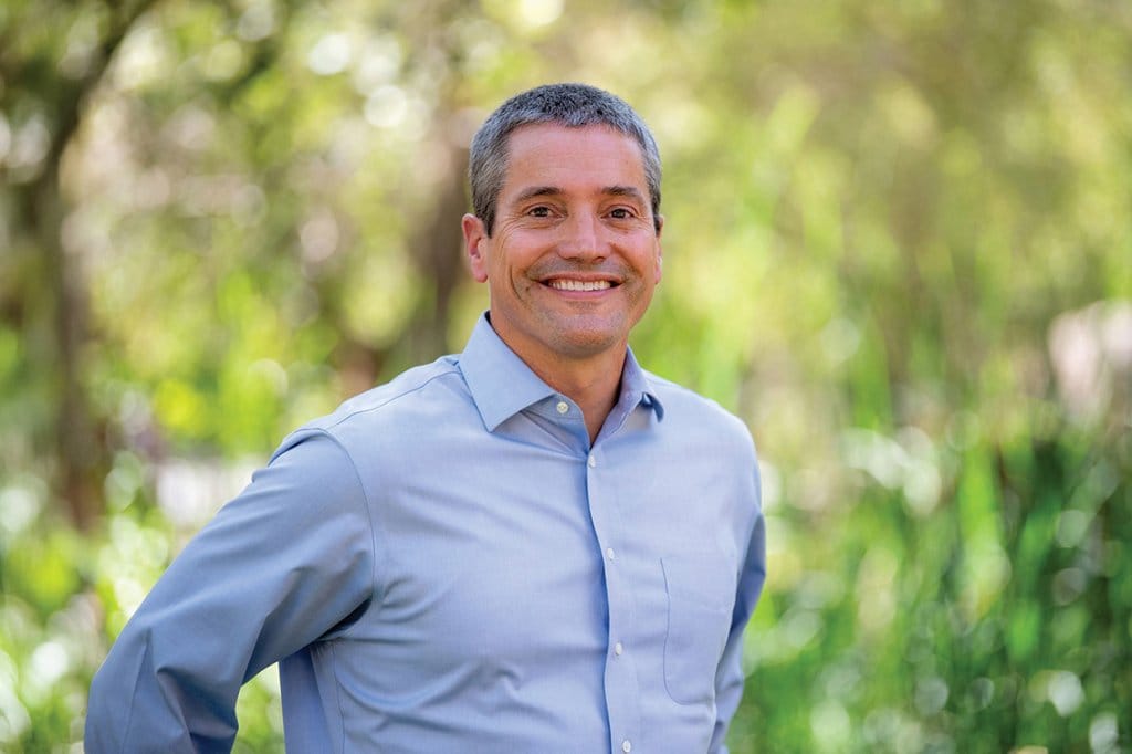 Wade Crowfoot, wearing a light blue button-up shirt, is standing outdoors with greenery and blurred trees in the background.