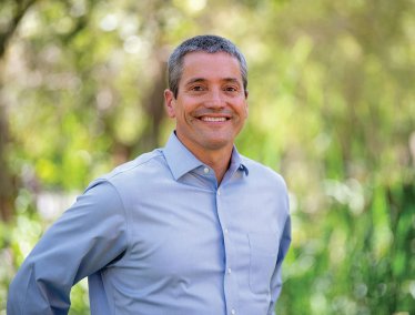 Wade Crowfoot, wearing a light blue button-up shirt, is standing outdoors with greenery and blurred trees in the background.