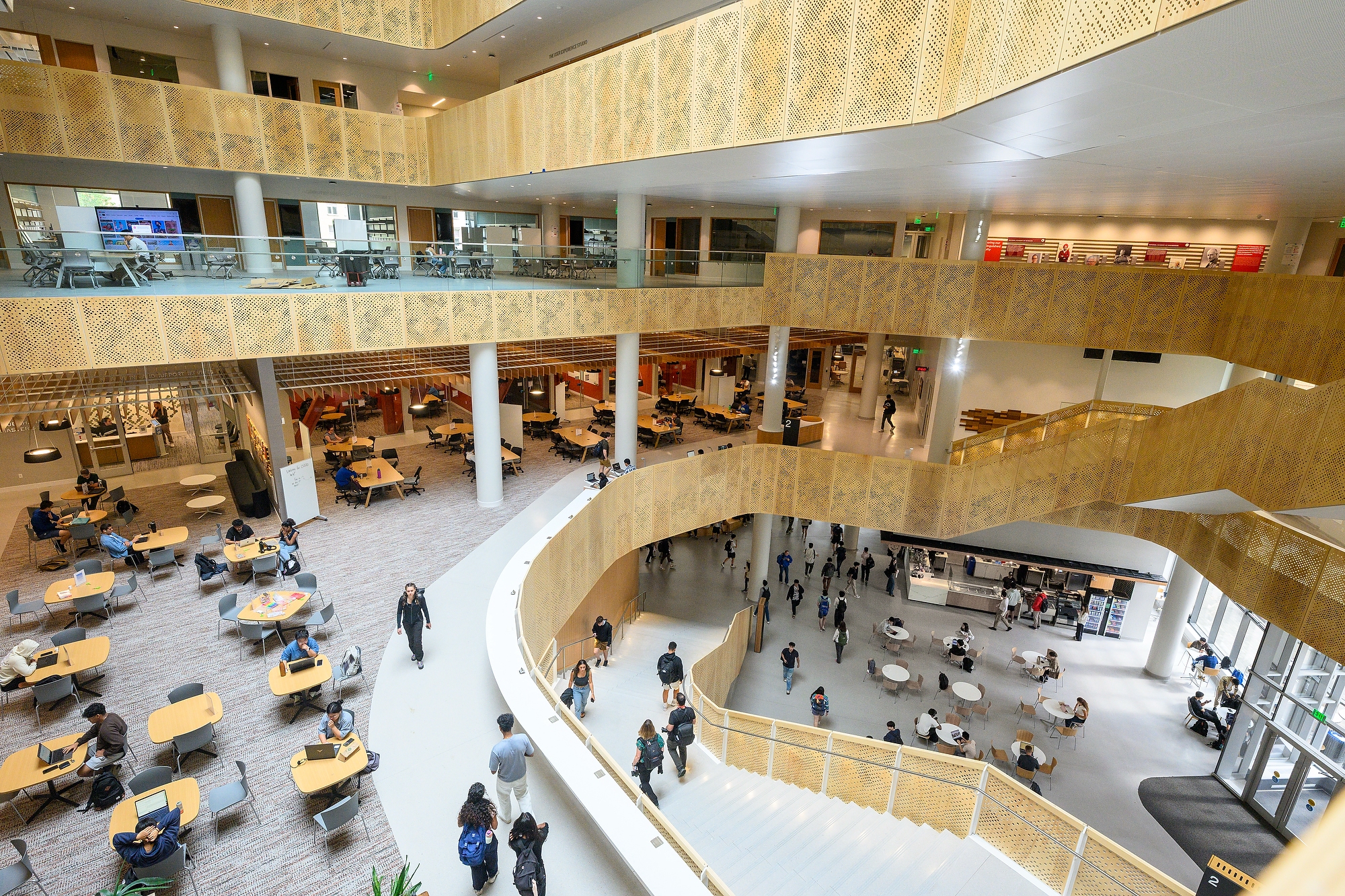 A spacious, multi-level modern building features open areas with tables and chairs, and people walking or sitting throughout the space.