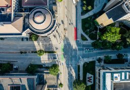An aerial view of an intersection in a campus area showing surrounding buildings, trees, and a landscaped area with a large red 'W' design.