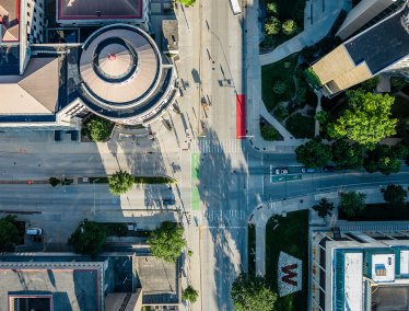 An aerial view of an intersection in a campus area showing surrounding buildings, trees, and a landscaped area with a large red 'W' design.