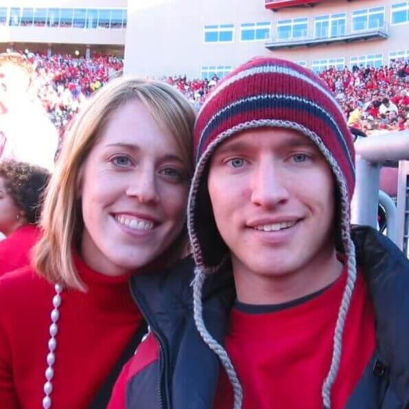 Two college-aged people dressed in red clothing are at a crowded St. Randall stadium, with one wearing a striped knit hat and the other wearing a necklace of large white beads.