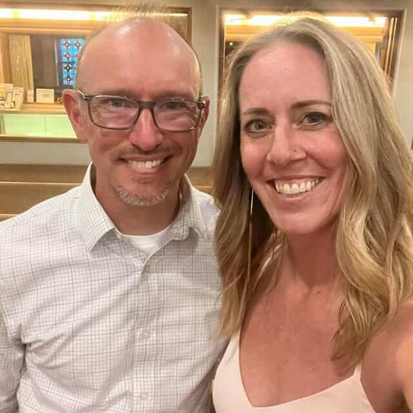 A couple standing indoors in a well-lit room, one wearing a light checkered shirt and the other in a sleeveless light-colored top.