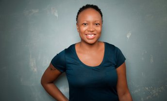 Kashana Cauley, wearing a dark blue short-sleeve shirt, is standing against a textured gray wall with one arm bent and resting on the hip.