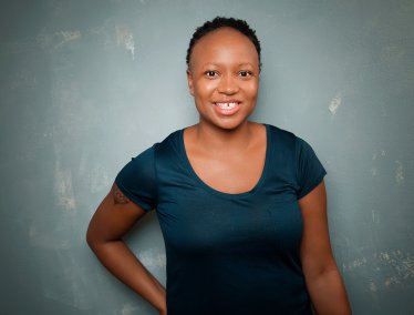 Kashana Cauley, wearing a dark blue short-sleeve shirt, is standing against a textured gray wall with one arm bent and resting on the hip.