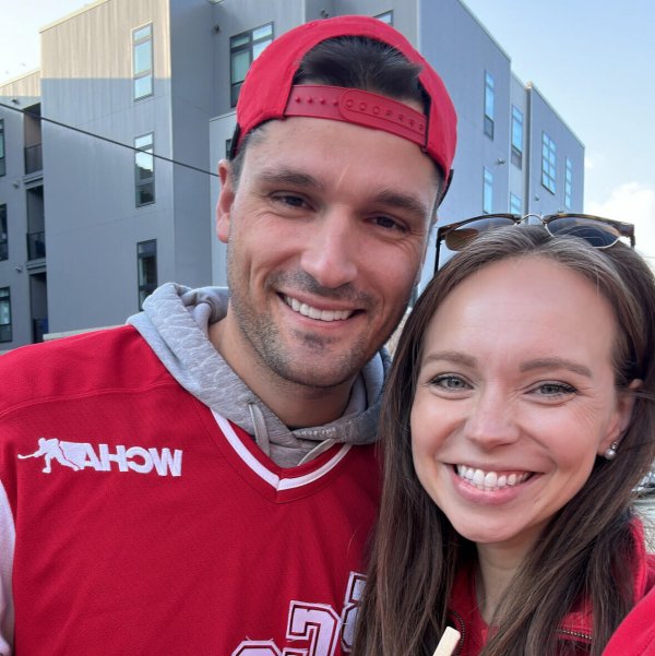 Two people wearing red sports jerseys pose together outdoors in front of modern gray buildings.