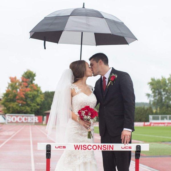 A couple dressed in wedding attire shares a kiss under a large black-and-white umbrella while standing on a track near a hurdle labeled 'Wisconsin.'
