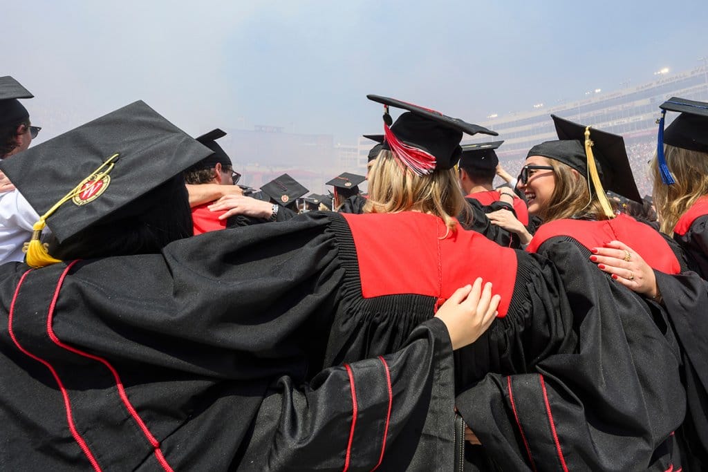 A group of UW graduates in black gowns with red accents stand arm-in-arm at Commencement in Camp Randall Stadium as they celebrate their achievement together.