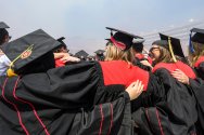 A group of UW graduates in black gowns with red accents stand arm-in-arm at Commencement in Camp Randall Stadium as they celebrate their achievement together.