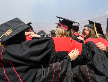 A group of UW graduates in black gowns with red accents stand arm-in-arm at Commencement in Camp Randall Stadium as they celebrate their achievement together.