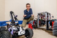 Dan Negrut kneels on an indoor sandy surface surrounded by three robotic vehicles with distinct mechanical components and wheel designs.