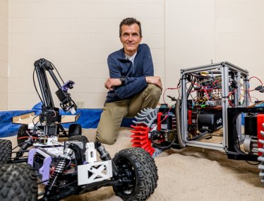 Dan Negrut kneels on an indoor sandy surface surrounded by three robotic vehicles with distinct mechanical components and wheel designs.