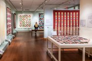 A gallery showcases colorful quilts on the walls and in a glass case, while a person sits on a bench observing one of the quilts.