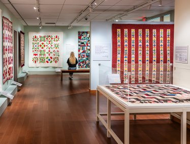 A gallery showcases colorful quilts on the walls and in a glass case, while a person sits on a bench observing one of the quilts.