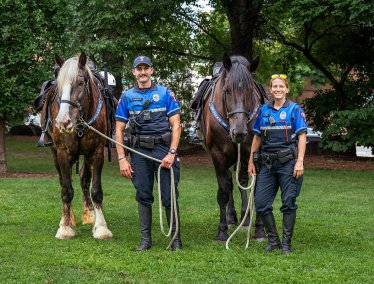 Two police officers stand in a grassy area with trees, holding the reins of two large horses.