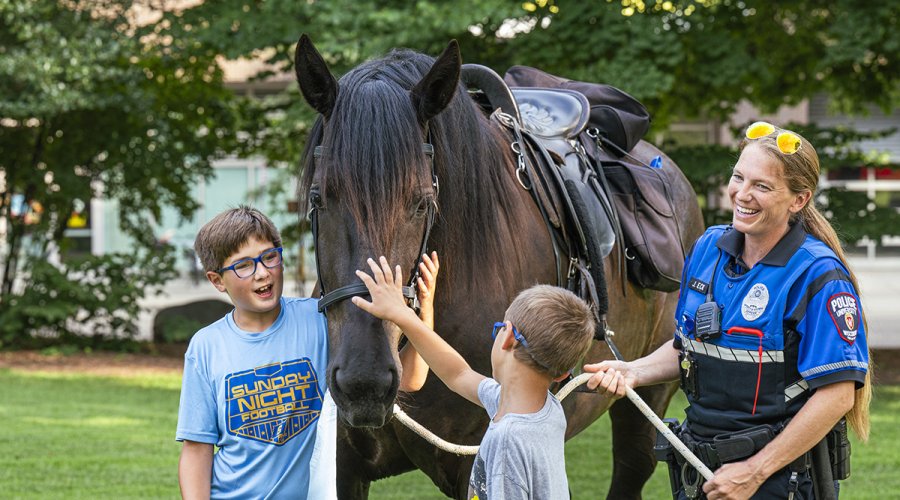 Two children reach out to pet a dark horse while a mounted police officer holds the reins in a grassy park.