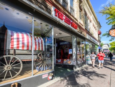 The front of an establishment with large glass windows displaying a red-and-white striped wagon, with the sign “The Red Shed” above the entrance and pedestrians walking along a sunny sidewalk.