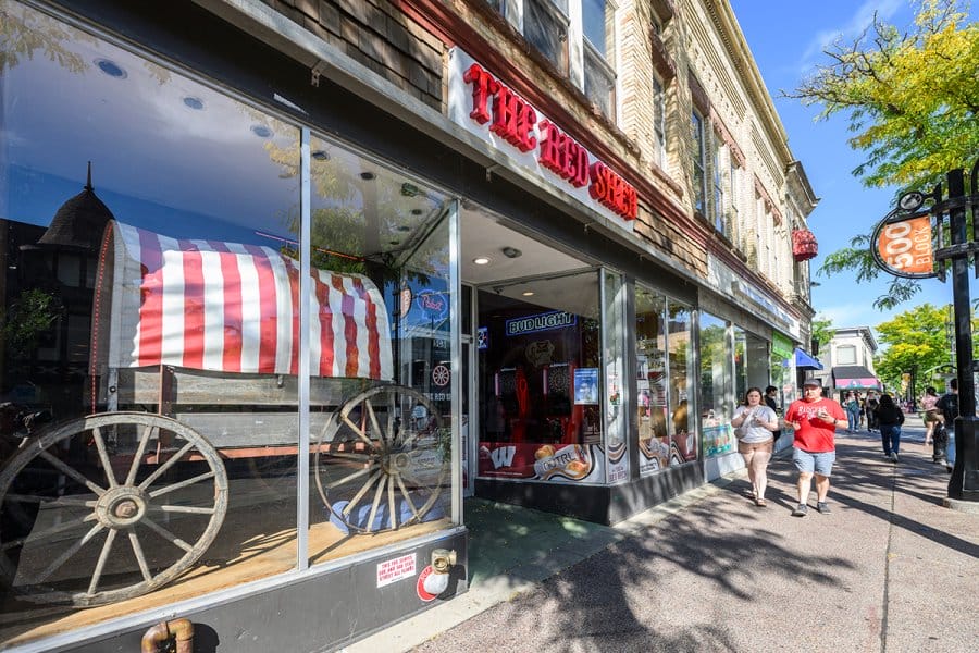 The front of an establishment with large glass windows displaying a red-and-white striped wagon, with the sign “The Red Shed” above the entrance and pedestrians walking along a sunny sidewalk.