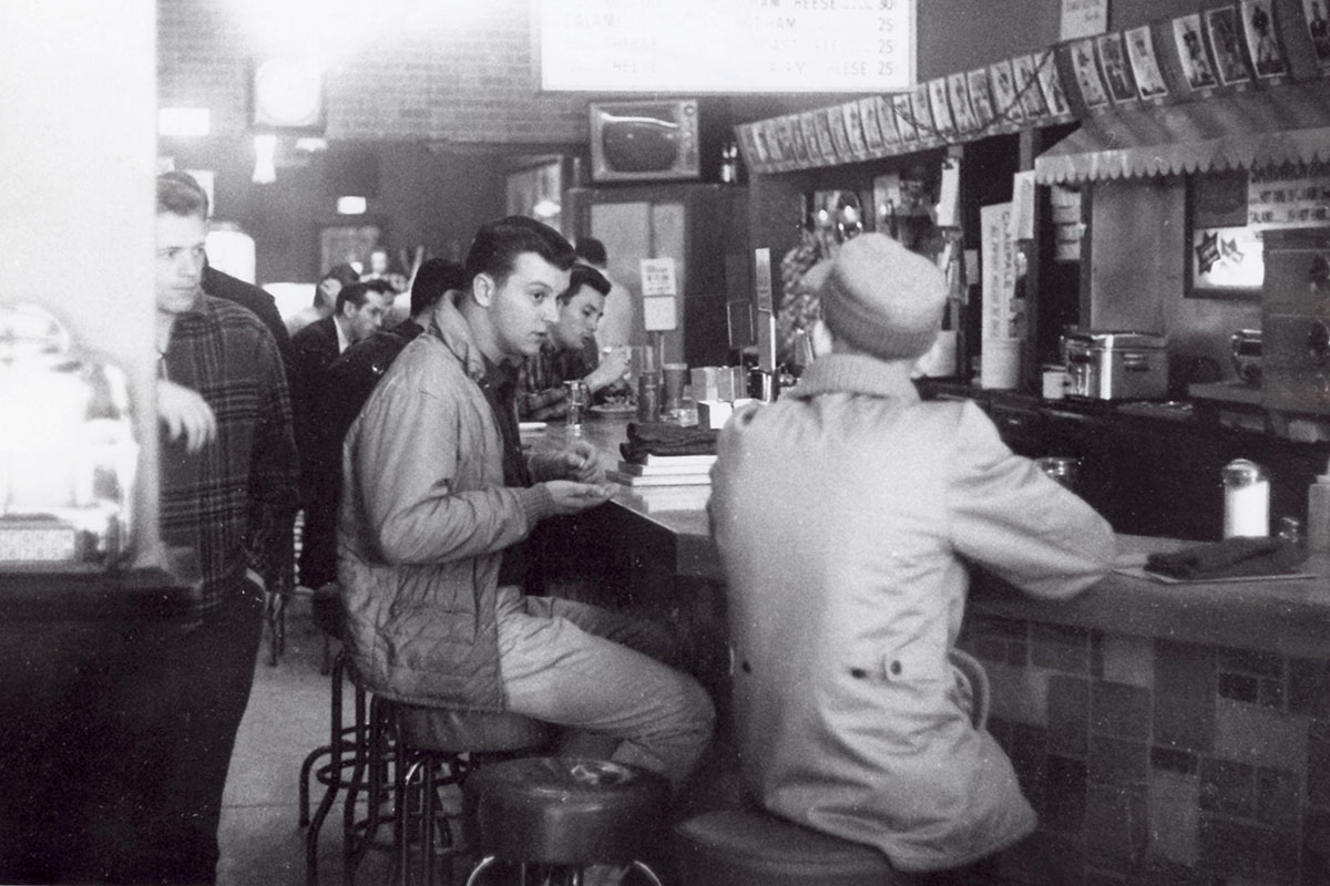 Grayscale image of individuals sitting at a diner counter, eating and conversing, with a menu board listing items like hamburgers and cheeseburgers overhead.
