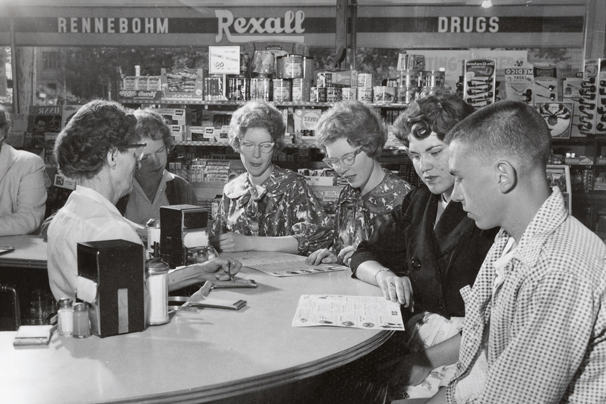 Grayscale image of people seated at a counter inside a drugstore with shelves of products and signage for 'Rennebohm' and 'Rexall Drugs.'