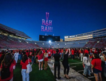 A large crowd of people, mostly dressed in red, gather on the football field at Camp Randall at night with a light display in the sky reading 'U RAH RAH!'