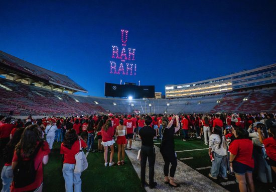 A large crowd of people, mostly dressed in red, gather on the football field at Camp Randall at night with a light display in the sky reading 'U RAH RAH!'