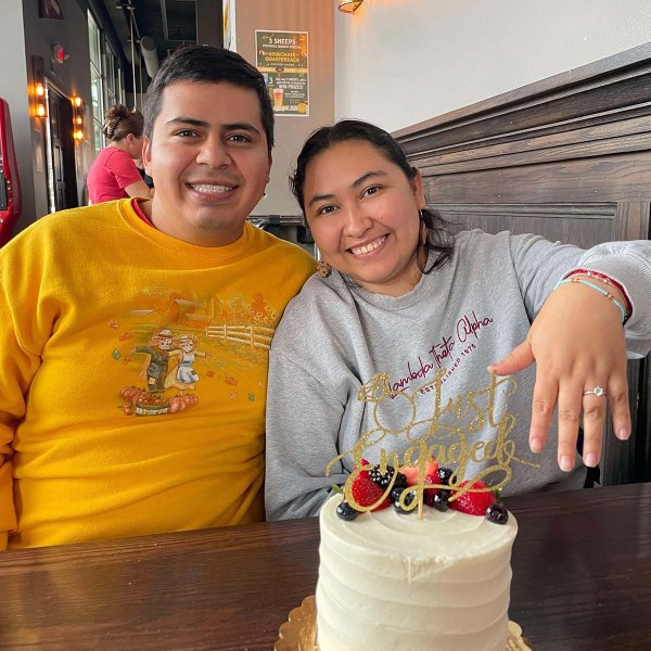 A couple sits at a restaurant table with a white cake topped with fruit and a “Just Engaged” decoration. The woman holds out her hand to show an engagement ring.