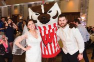 A bride and groom in wedding attire pose with Bucky Badger at an indoor reception.