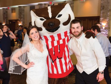 A bride and groom in wedding attire pose with Bucky Badger at an indoor reception.