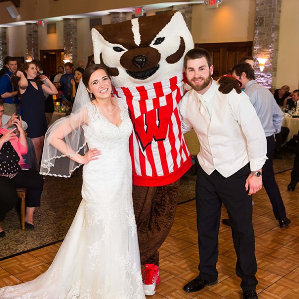 A bride and groom in wedding attire pose with Bucky Badger at an indoor reception.