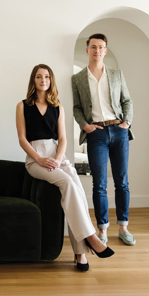 Grace and Michael van Meurer are posed indoors in a modern, minimalist setting, with one seated on a dark sofa and the other standing near an arched doorway.
