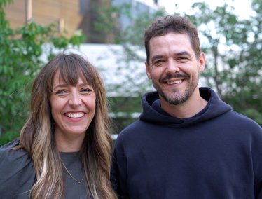 Meagan Hahn and Shawn Wiederhoeft stand outdoors smiling at the camera, with greenery and buildings in the background.