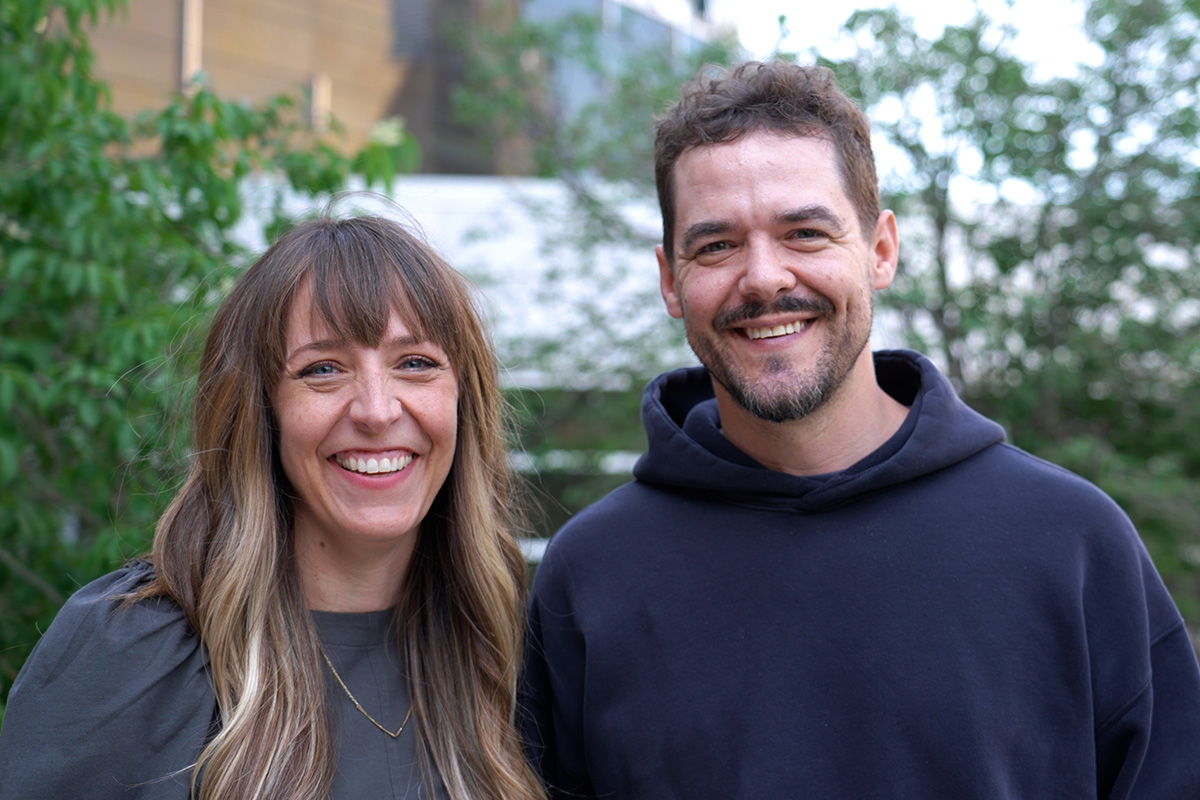 Meagan Hahn and Shawn Wiederhoeft stand outdoors smiling at the camera, with greenery and buildings in the background.