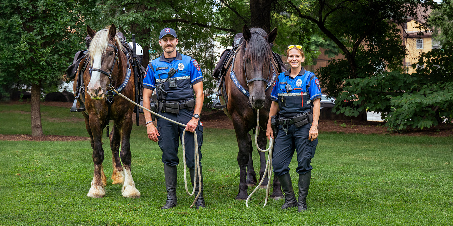 Two police officers stand in a grassy area with trees, holding the reins of two large horses.