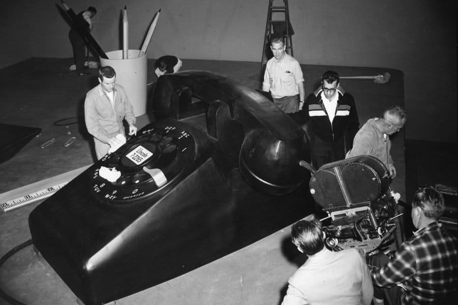 Grayscale photo of a group of people working around an oversized rotary telephone prop on a studio set.