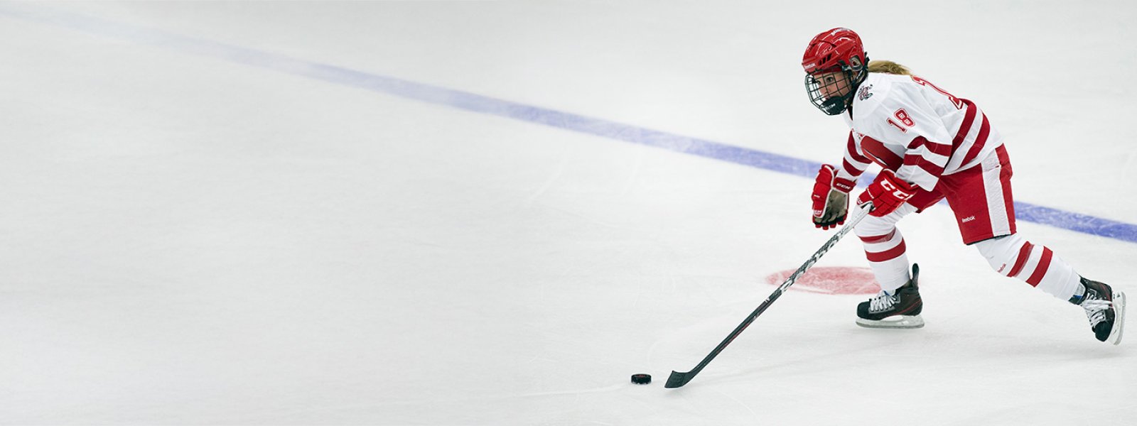 Ice hockey player in a red and white uniform skating on the rink while controlling the puck with a stick.