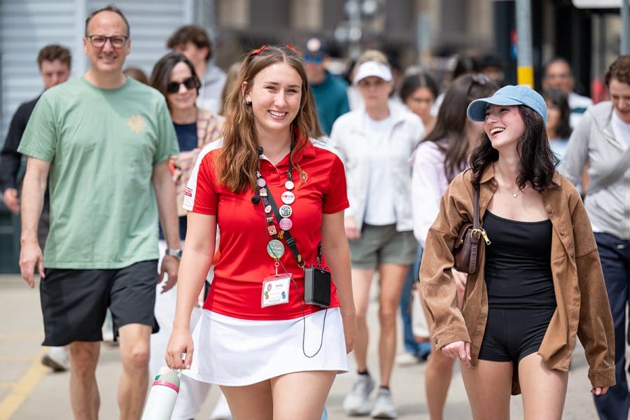 A group of people walking outdoors, with a person in a red shirt and white skirt wearing a lanyard and buttons leading the group.