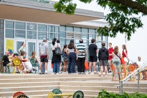 A group of people stands outside the Wisconsin Union Theater building, listening to two tour guides in red shirts .