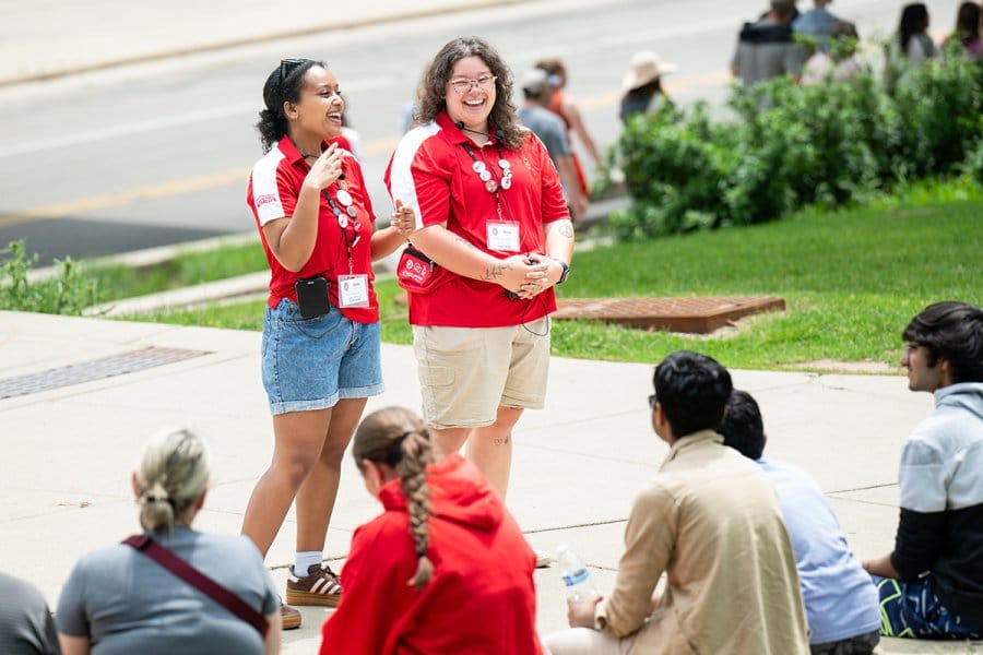 Two tour guides wearing red University of Wisconsin polo shirts and name badges stand on a sidewalk speaking to a seated group of people outdoors near greenery.