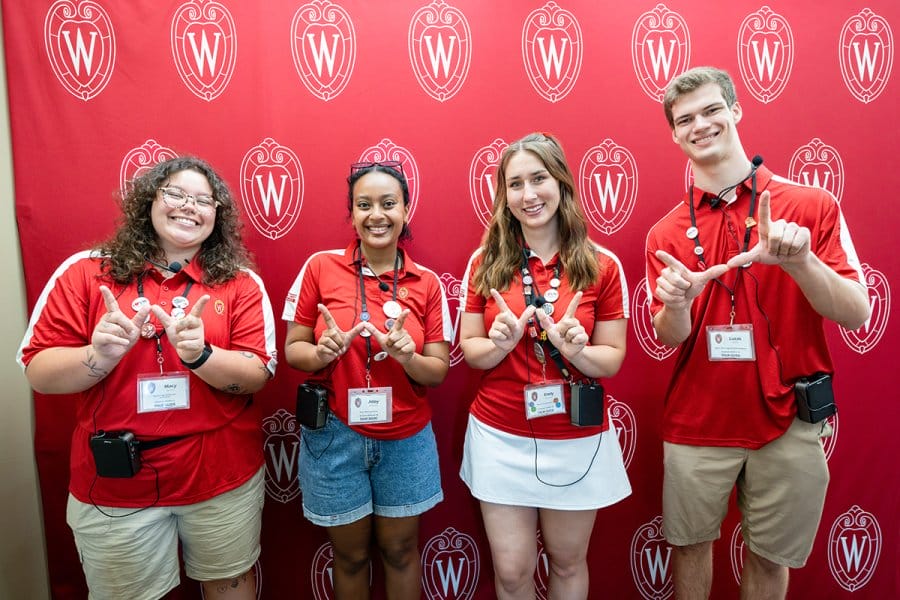 Four campus tour guides wearing red University of Wisconsin polo shirts and name badges stand in front of a red backdrop with the university’s crest, each making a 'W' hand gesture.