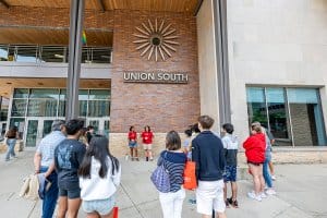 People stand in a group outside Union South, listening to two tour guides in red shirts near the building entrance.