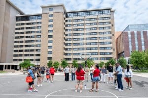 A tour group gathers on a basketball court in front of Sellery residence hall, with guides in red shirts speaking to them.